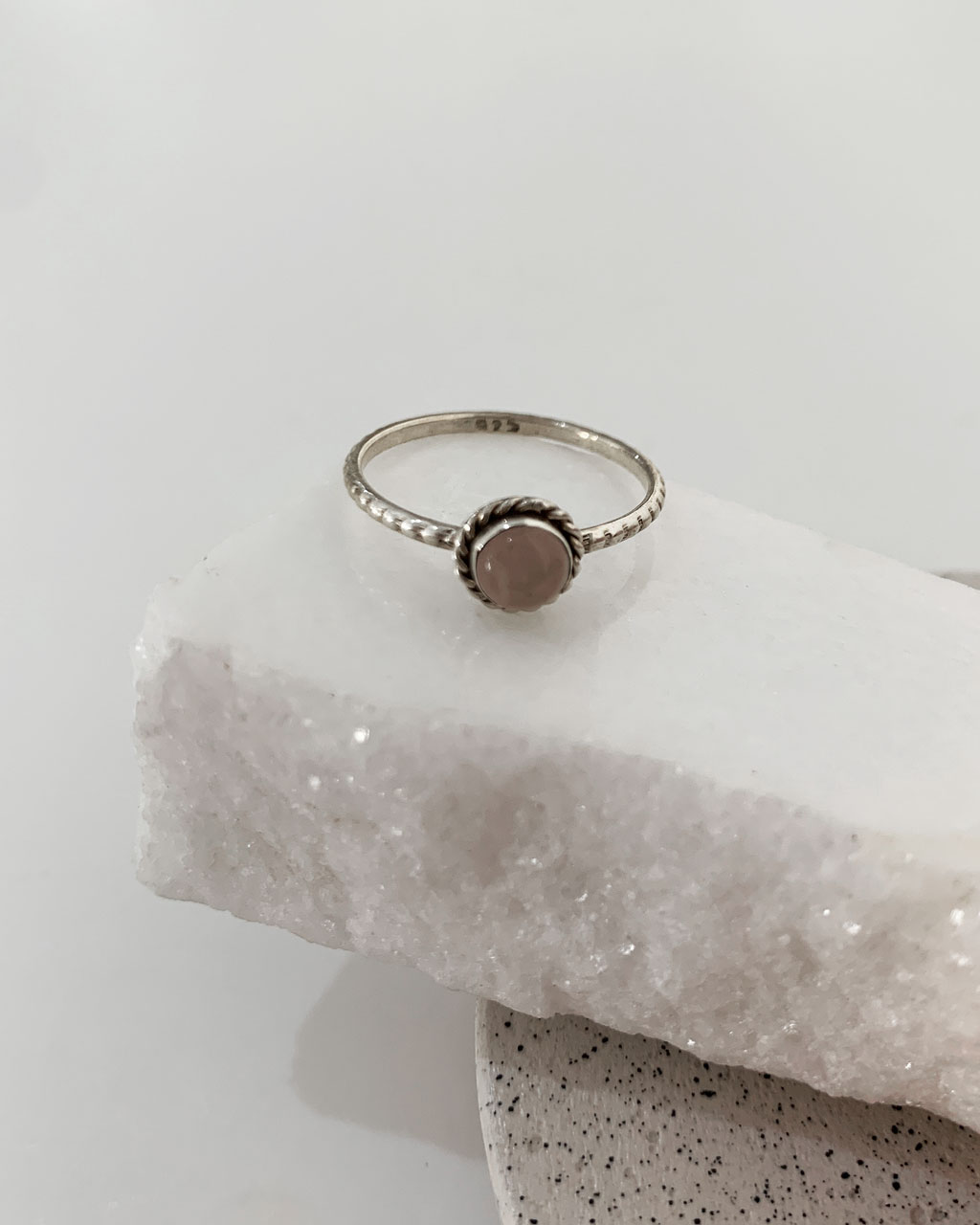 Close-up image of a shiny, sterile silver ring with a Rose quartz Stone on a white marble on a concrete coaster