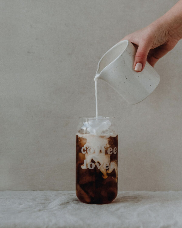 a close up of a person's hand pouring milk into A tall drinking glass with white engraved text saying coffee lovers. filled with an iced coffee