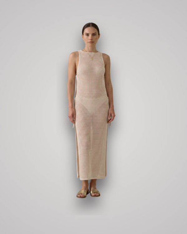 Woman standing in a beige sleeveless crochet maxi dress with side slits, paired with gold sandals against a light gray backdrop.